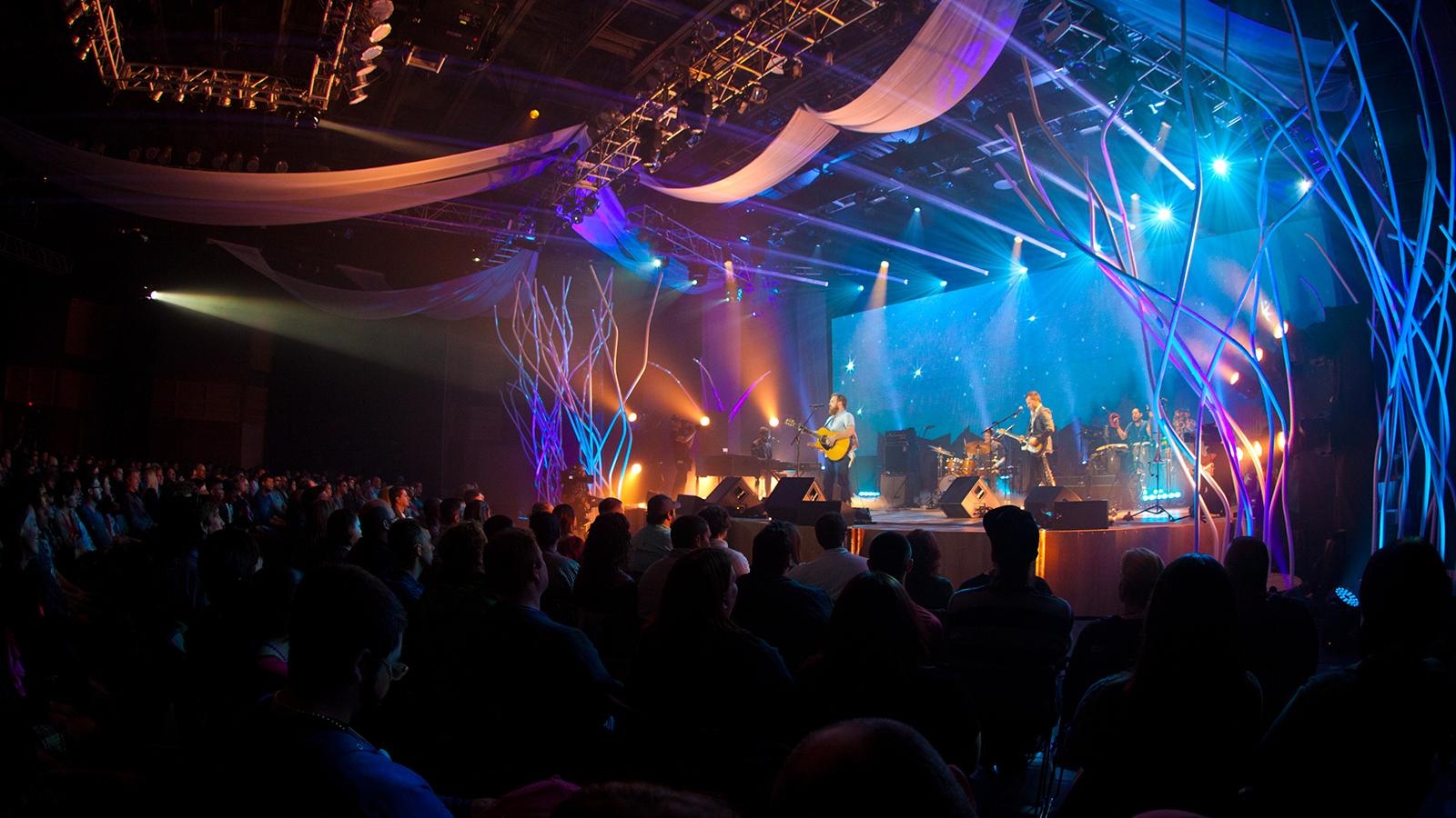 A band plays music in a large, dark auditorium with several bright blue and purple lights pointing at the stage.