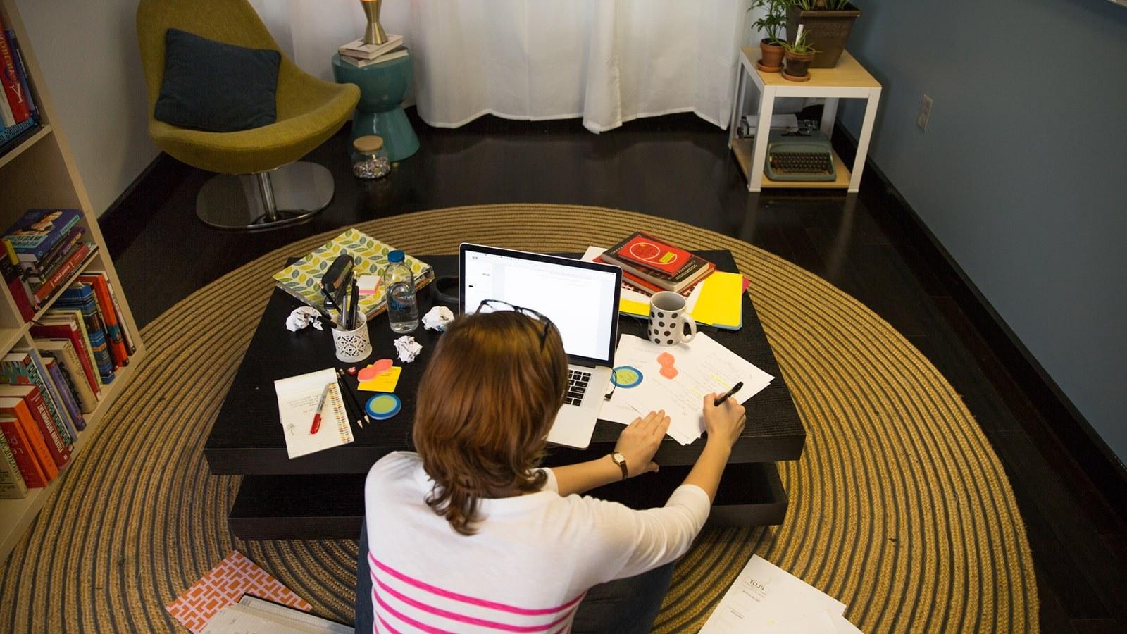 A student sits cross-legged on the floor in front of a coffee table with a laptop, 论文, notebooks, and pens.