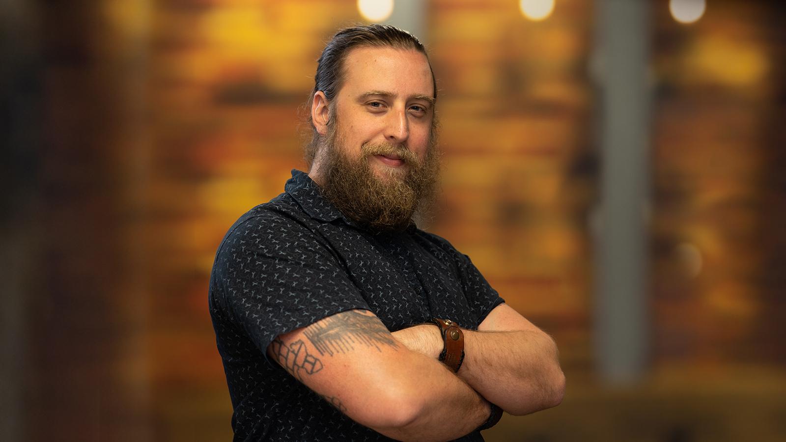 Kyle is standing with his arms crossed, smiling gently. He is wearing a short sleeve blue shirt and his hair is in a bun.