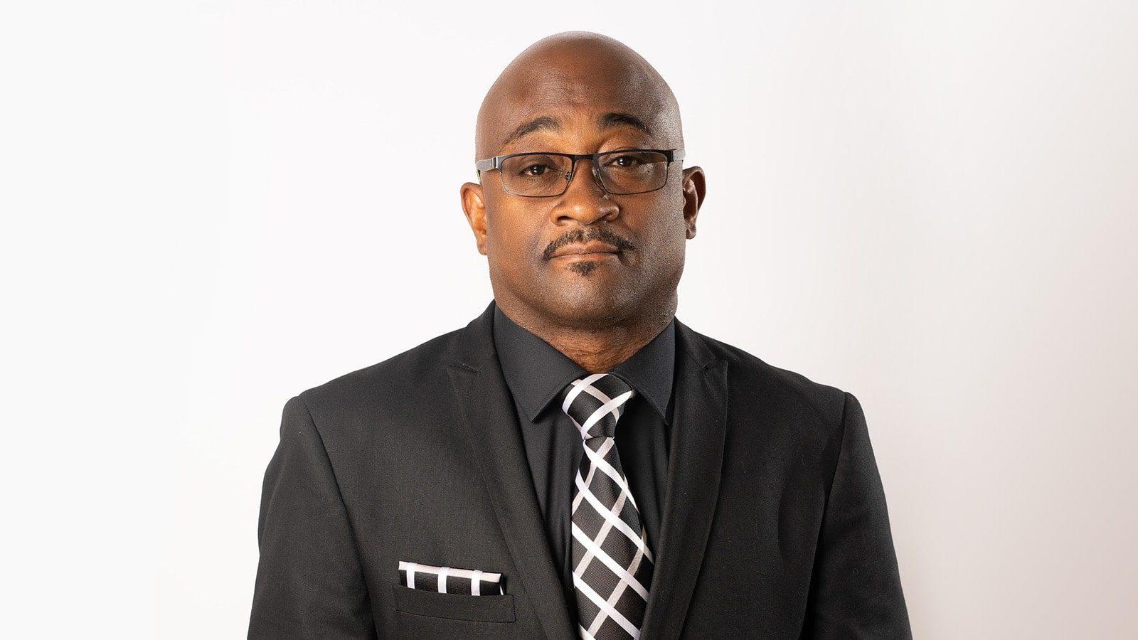 A headshot of Christopher Rapley wearing glasses and an all-black suit with a black-and-white tie and pocket square.