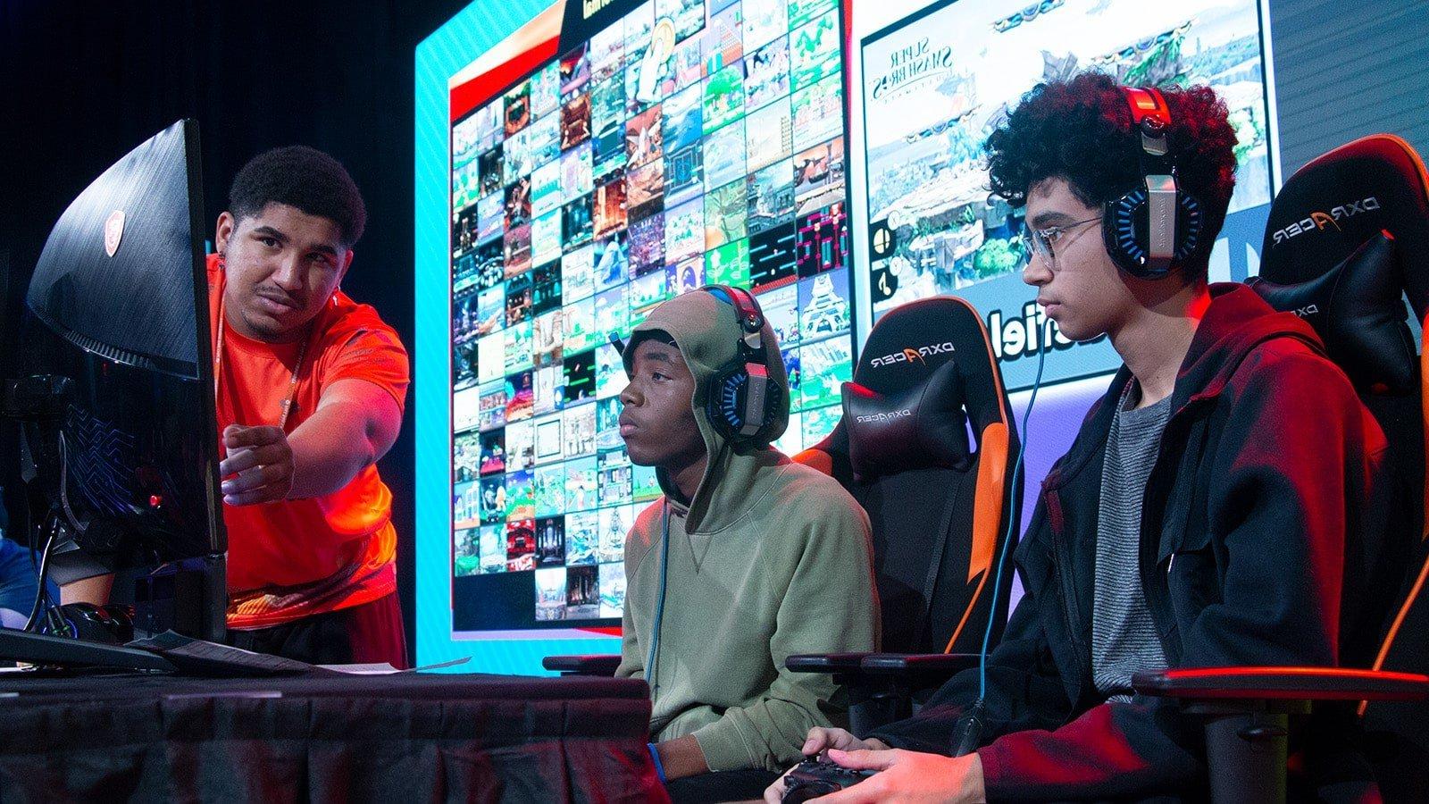 Two Florida high school students seated on stage in front of a gaming monitor with a 满帆 Armada athlete helping them to their right.