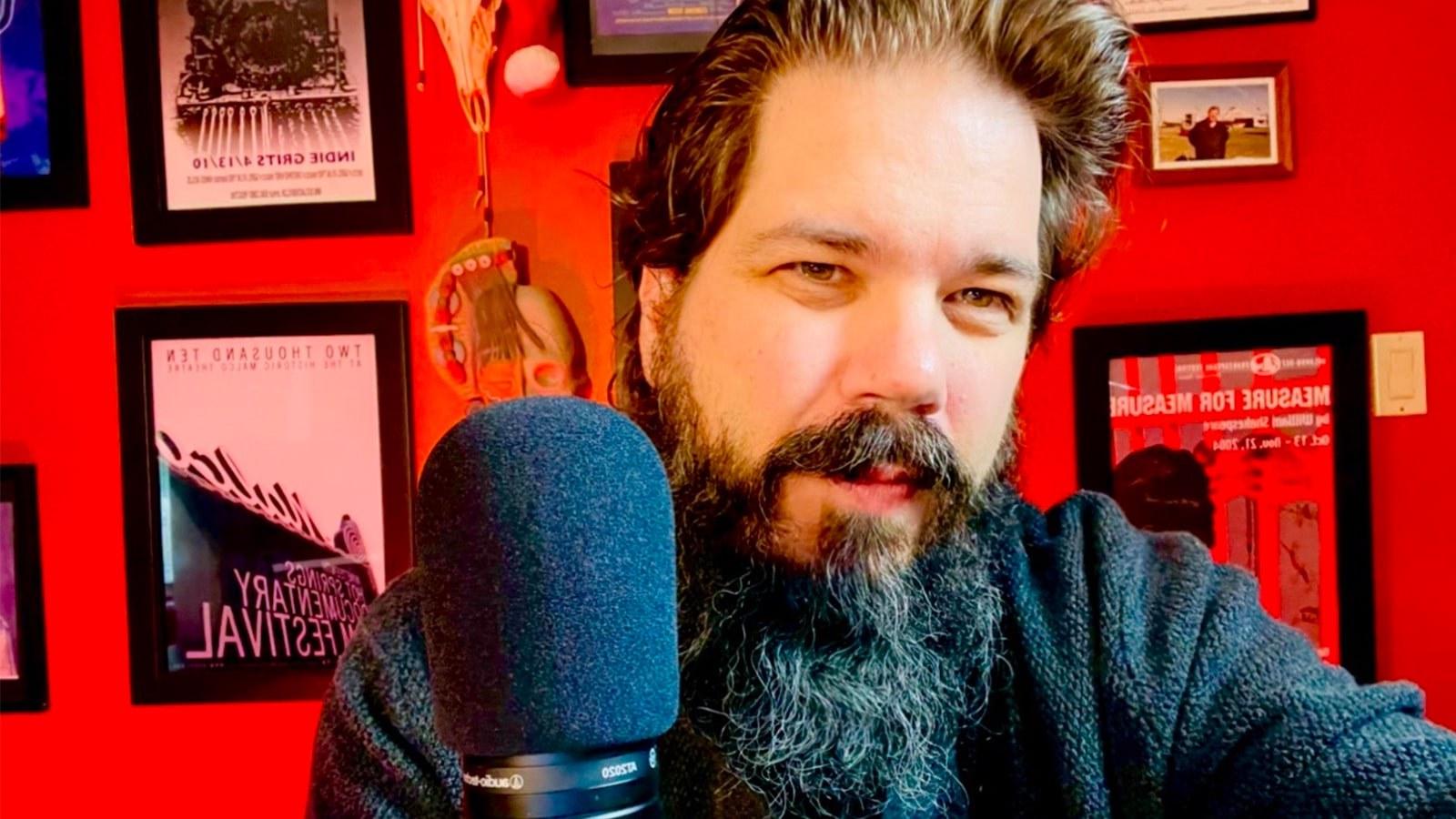 克里斯托弗·拉姆齐, a man with a long black and grey beard and short hair sitting with a microphone in front of a red wall filled with framed film posters.