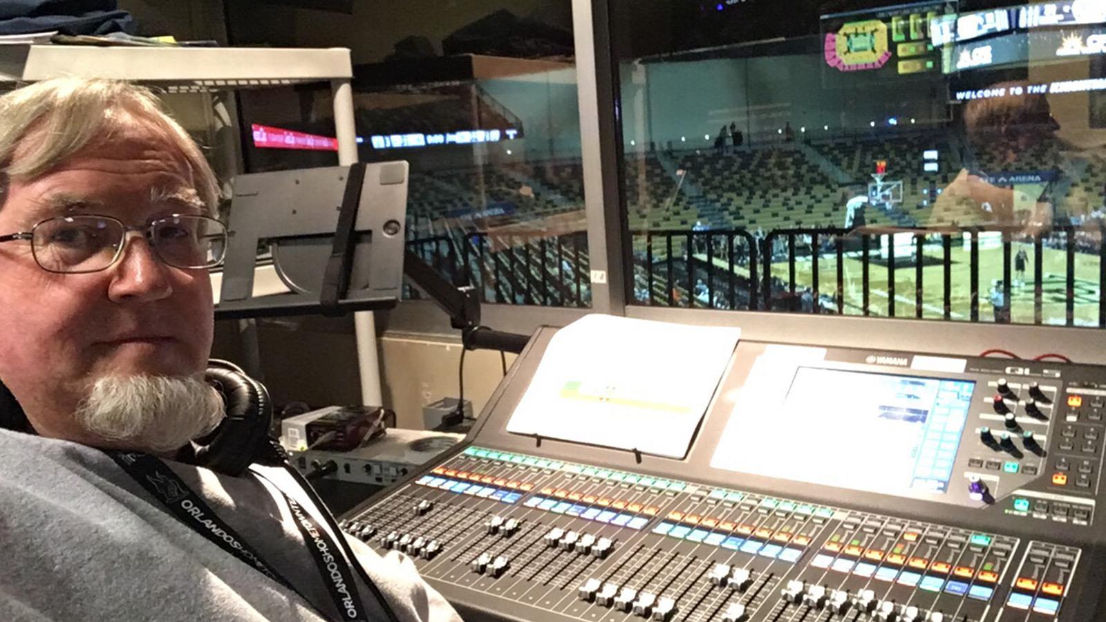 A man seated at an audio console overlooking a basketball court in a live arena.