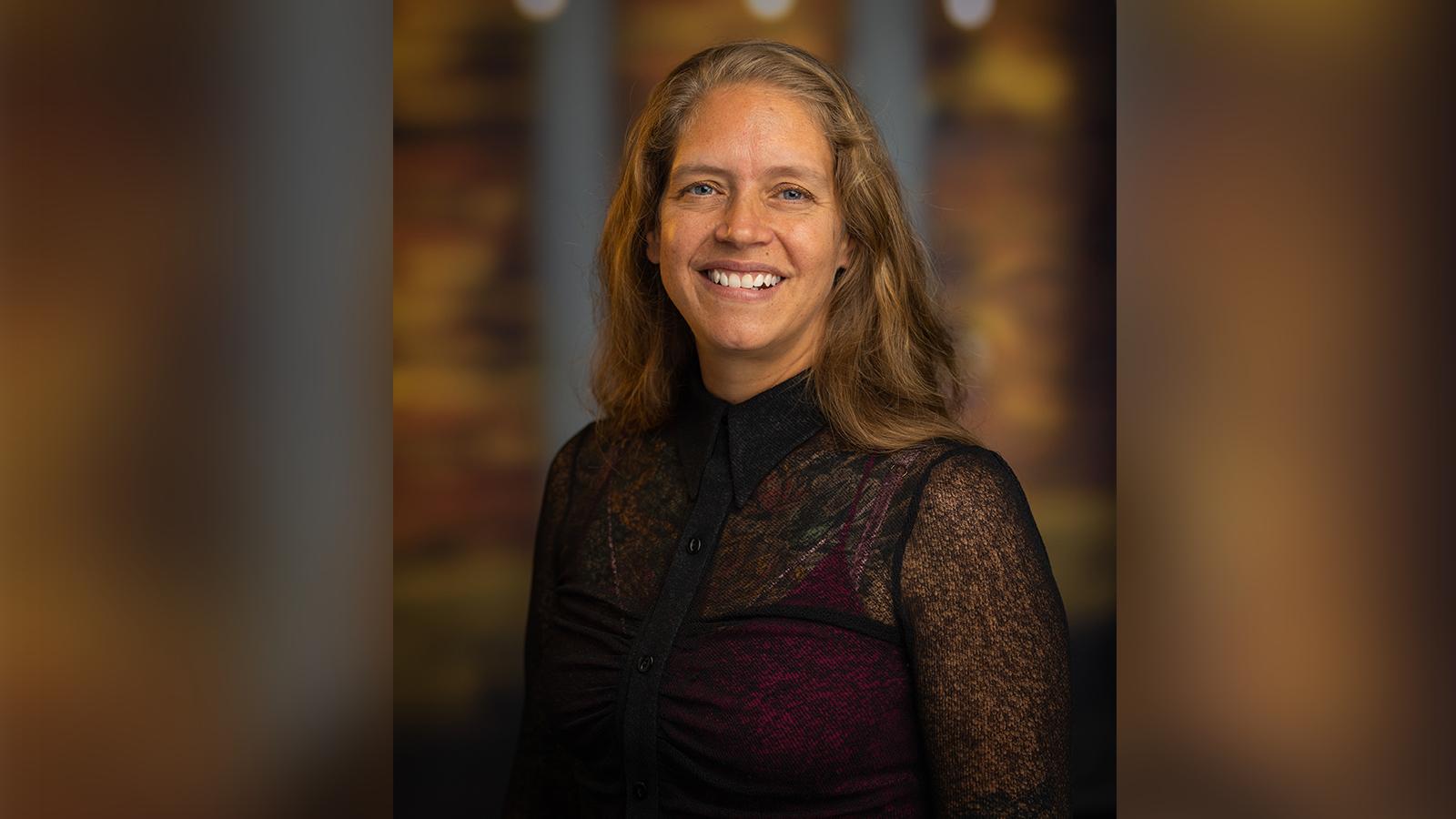 Phoebe Elefante, a woman with shoulder-length light brown hair and a black blouse smiling in front of an out-of-focus brown backdrop.