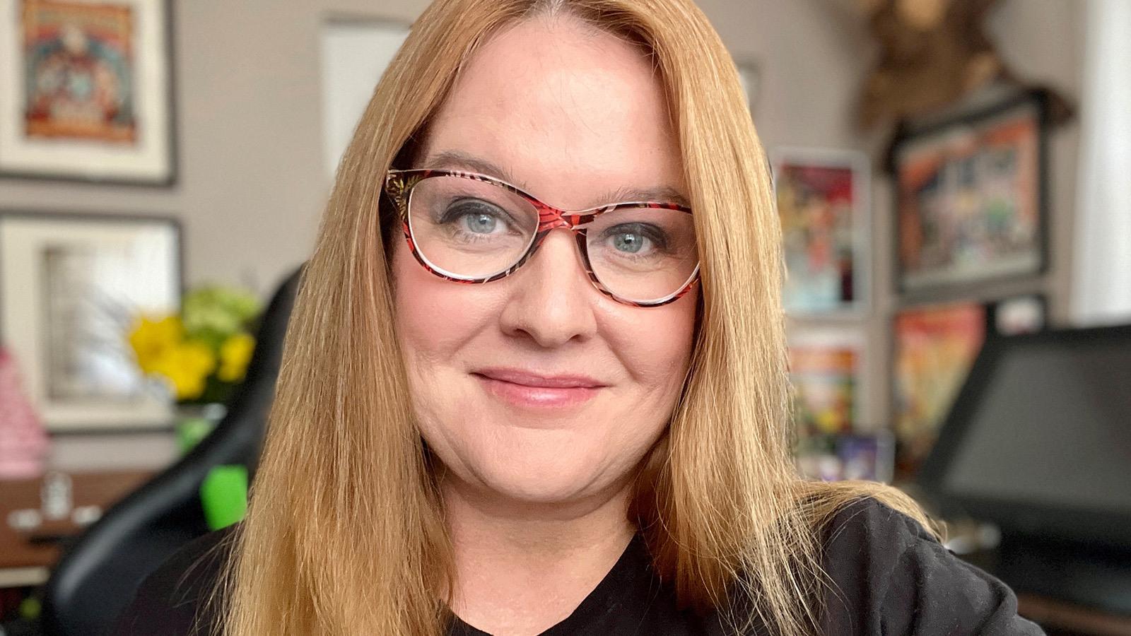 Amy Williams sits in her home design studio. The walls are covered in framed artwork. She is wearing glasses and smiling.