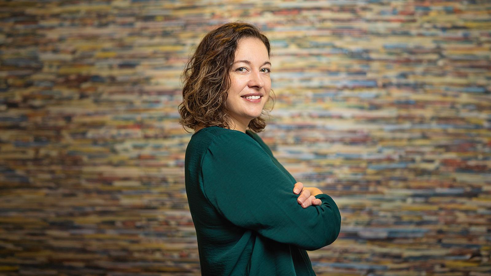 Aron Gelineau stands with her arms crossed and smiles at the camera. She has brown, curly hair and is wearing a teal sweater.