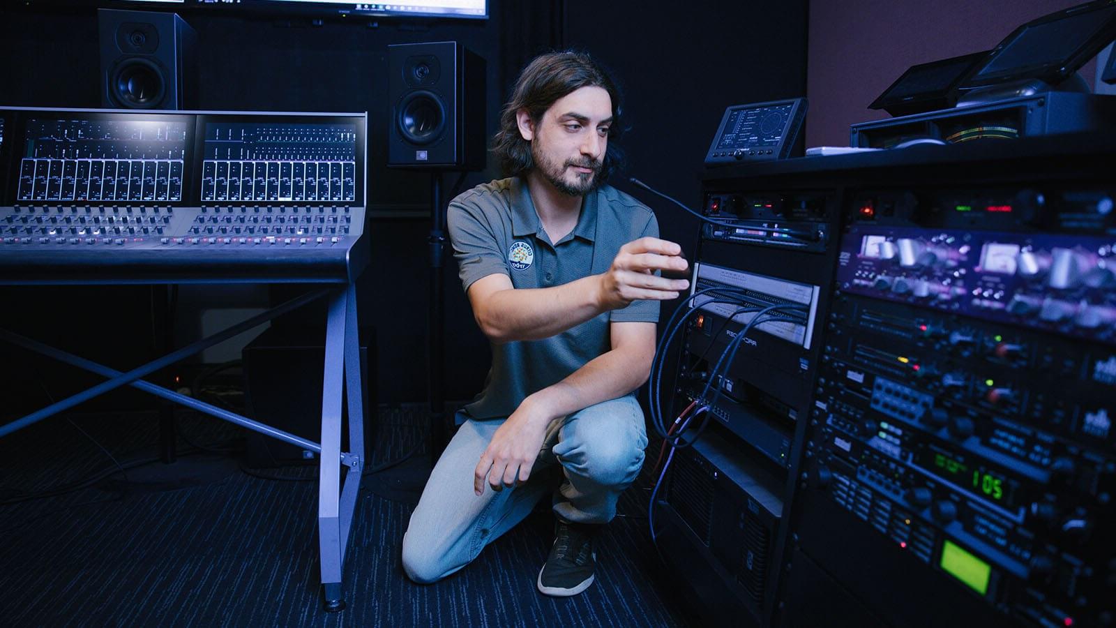 Dave Harris crouches next to broadcast equipment, including mixing consoles and a communications system.