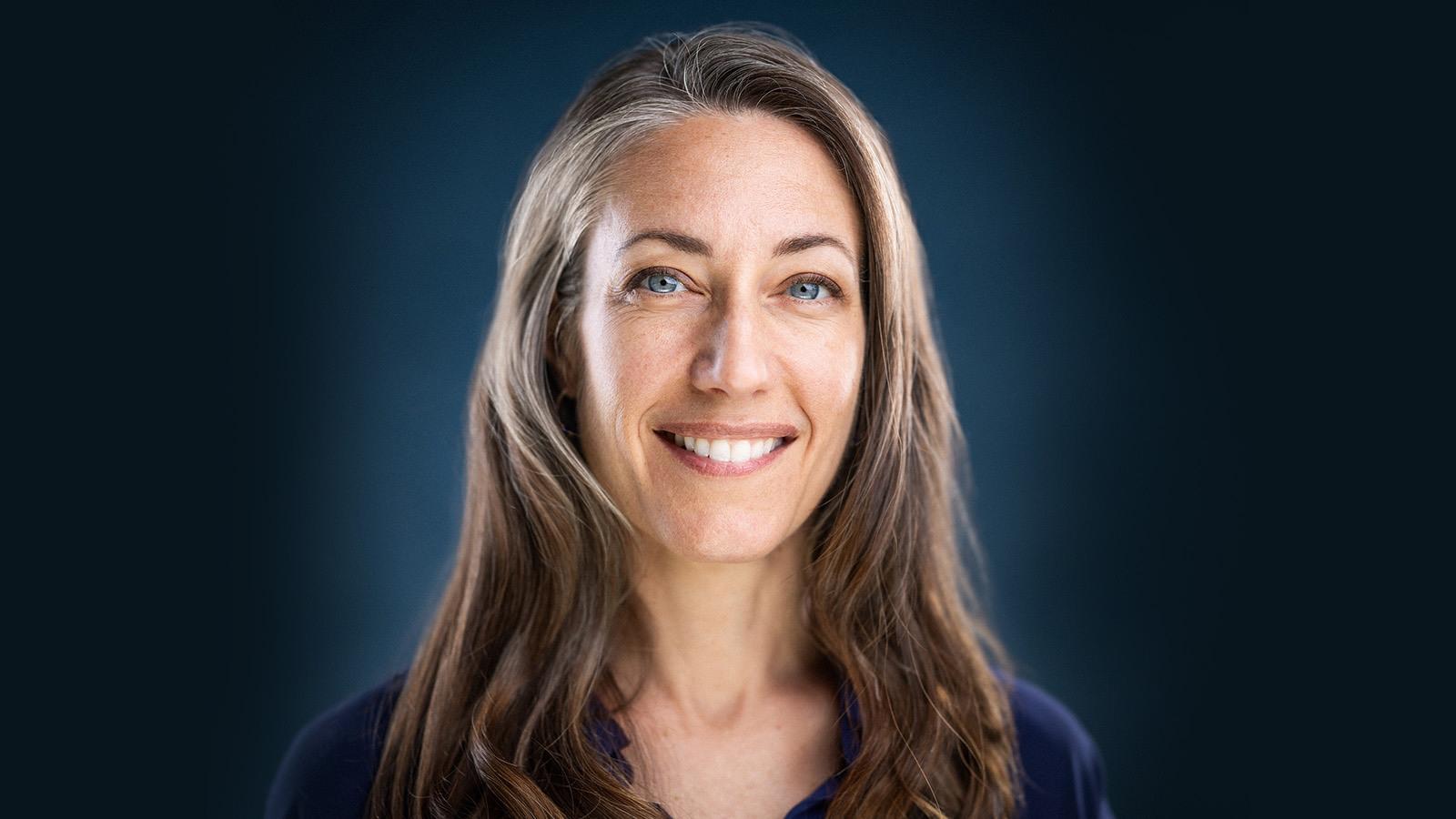 Kelly sits in front of a blue background wearing a blue blouse. She has blue eyes and is smiling.