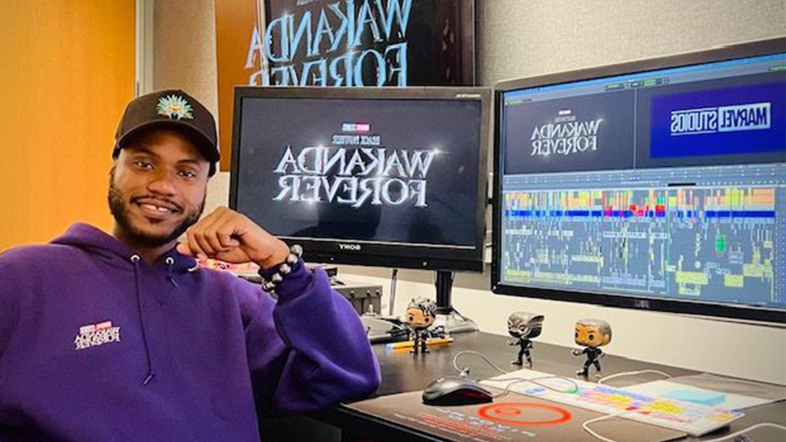 Alain sits at a desk with multiple monitors that read “Wakanda Forever” and “Marvel Studios.” He is wearing a purple hoodie.