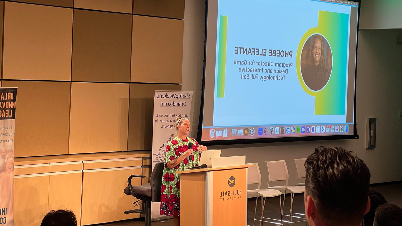 Phoebe Elefante stands at the podium speaking to the audience in the Full Sail venue. She is wearing a brightly colored print dress.