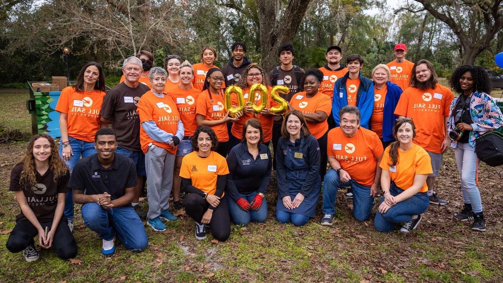 A group photo of students and staff wearing bright orange Full Sail University t-shirts. They are also holding golden balloons that spell 200.