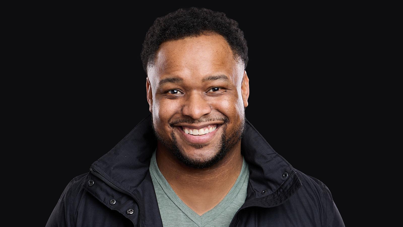 James Lewis smiles at the camera. He is wearing a black jacket and green v-neck t-shirt.