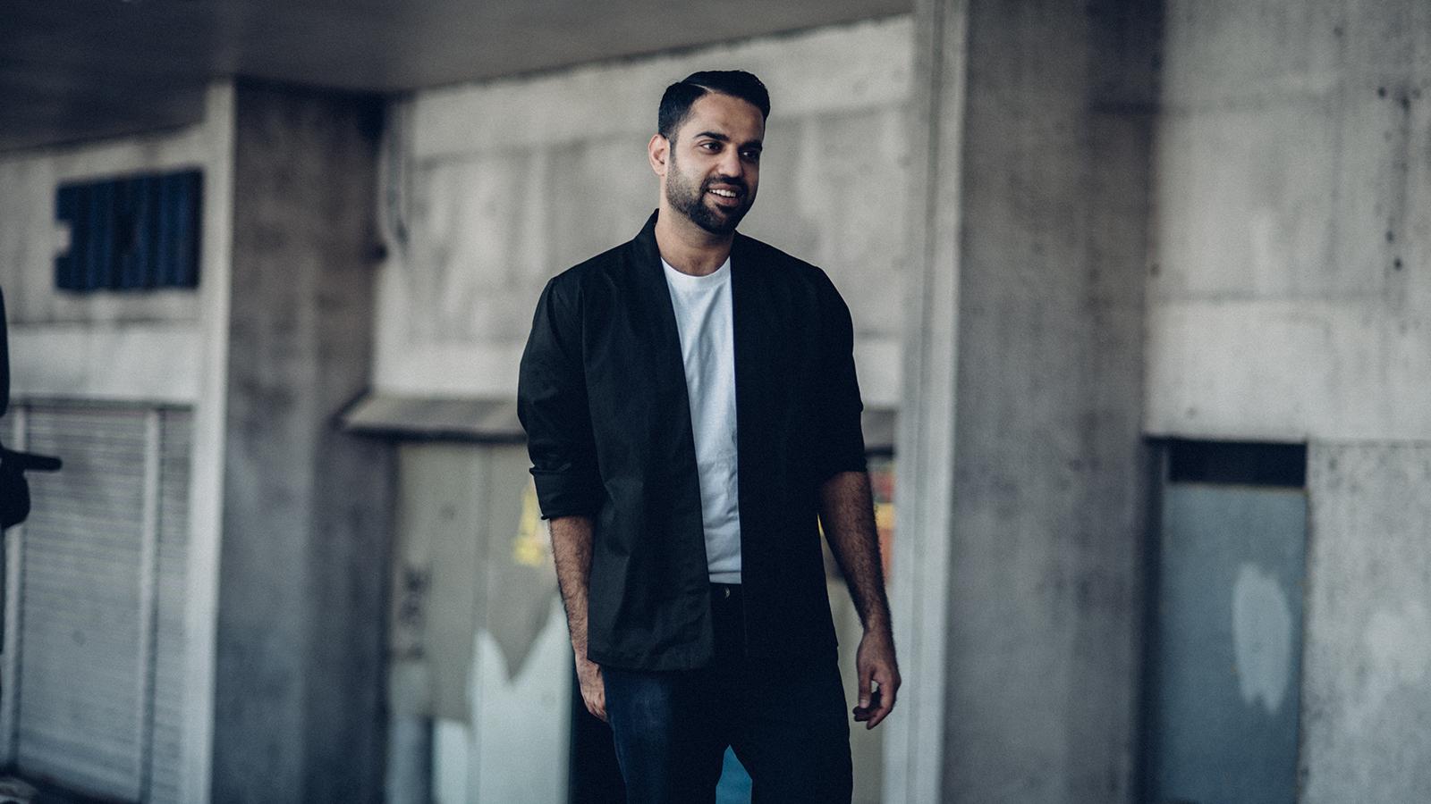 A well-dressed man in a white t-shirt and black blazer posing in front of a worn-down building.