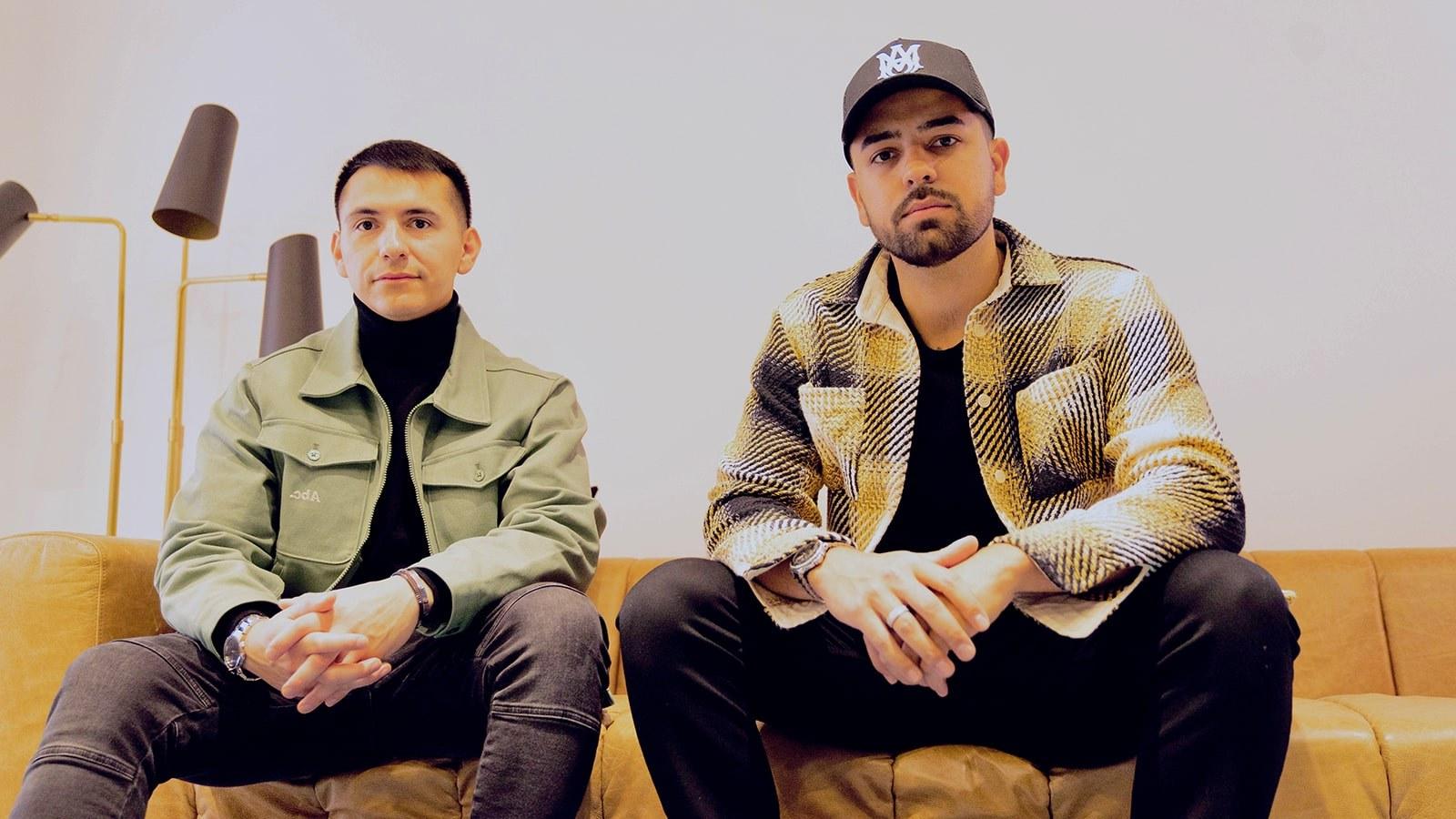 Two men seated side by side on a light brown leather couch against a white backdrop.