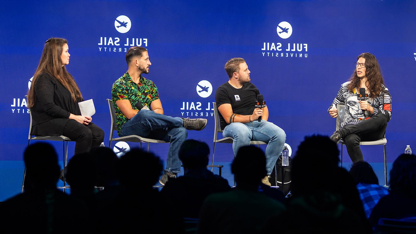 Four people seated on a stage, talking and holding microphones in front of a large crowd.