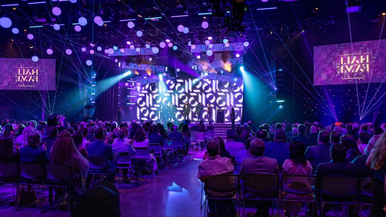 The 名人堂 2023 inductees st和 on a stage, surrounded by beautiful purple lighting in a large auditorium. 观众在鼓掌.