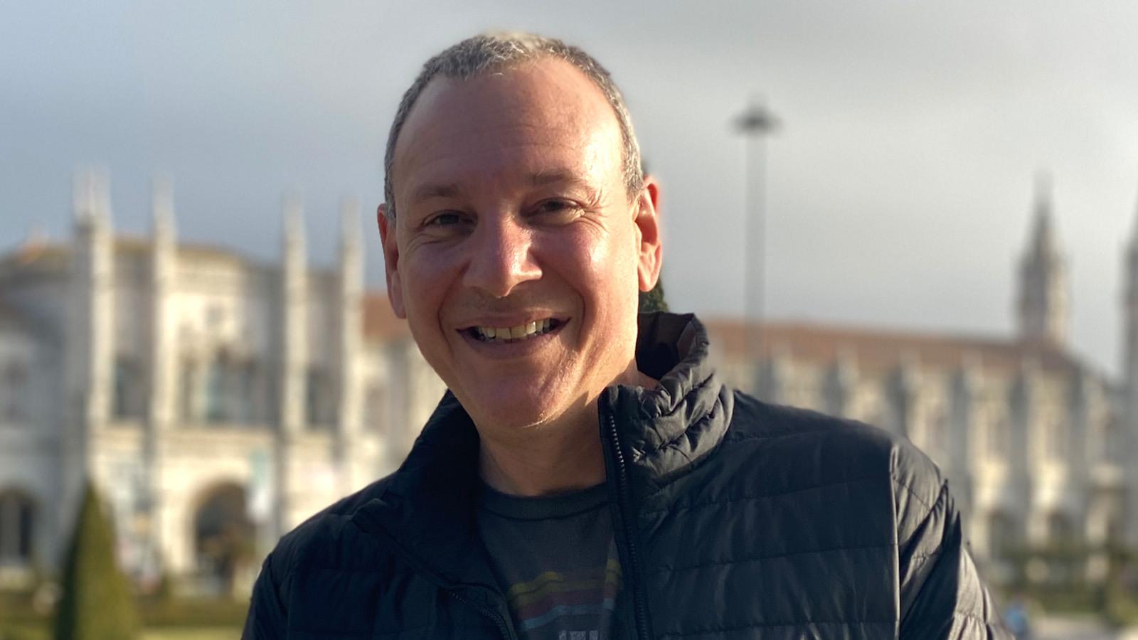 Sean Spuehler stands outdoors in front of a large building. He is smiling and wearing a black jacket and a blue t-shirt.