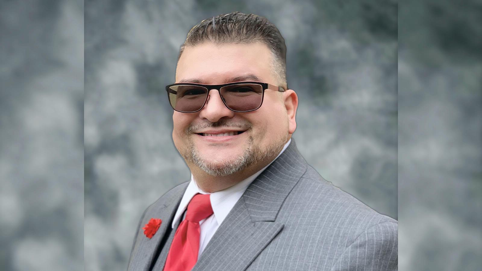 Will Torres wears a gray pinstriped suit with a red tie, a red carnation on the lapel, 和太阳镜.