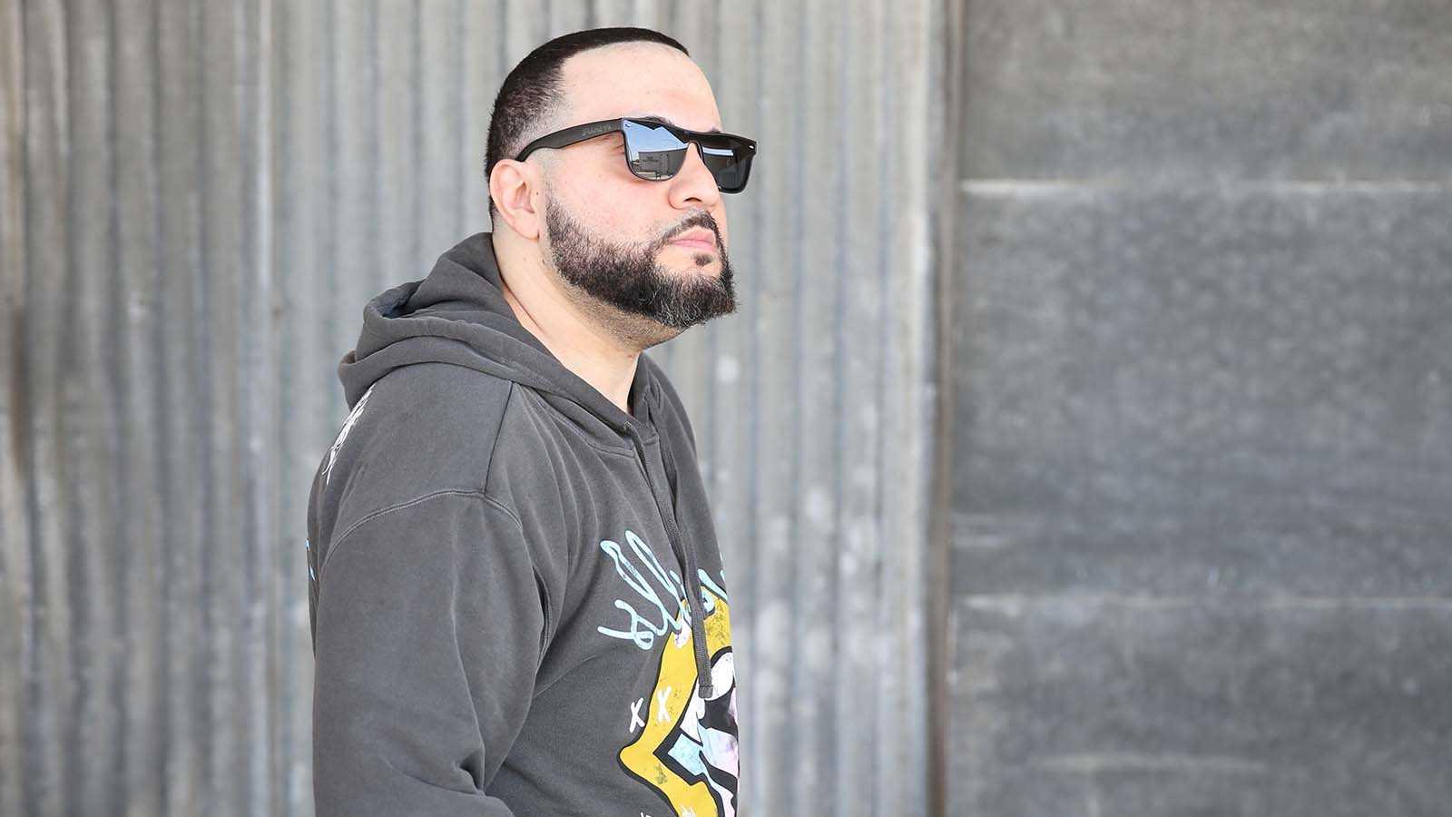 Full Sail grad George Doman wearing black sunglasses and a grey pullover hoodie with a yellow and light blue design, standing in front of a large stone wall while looking off into the distance.