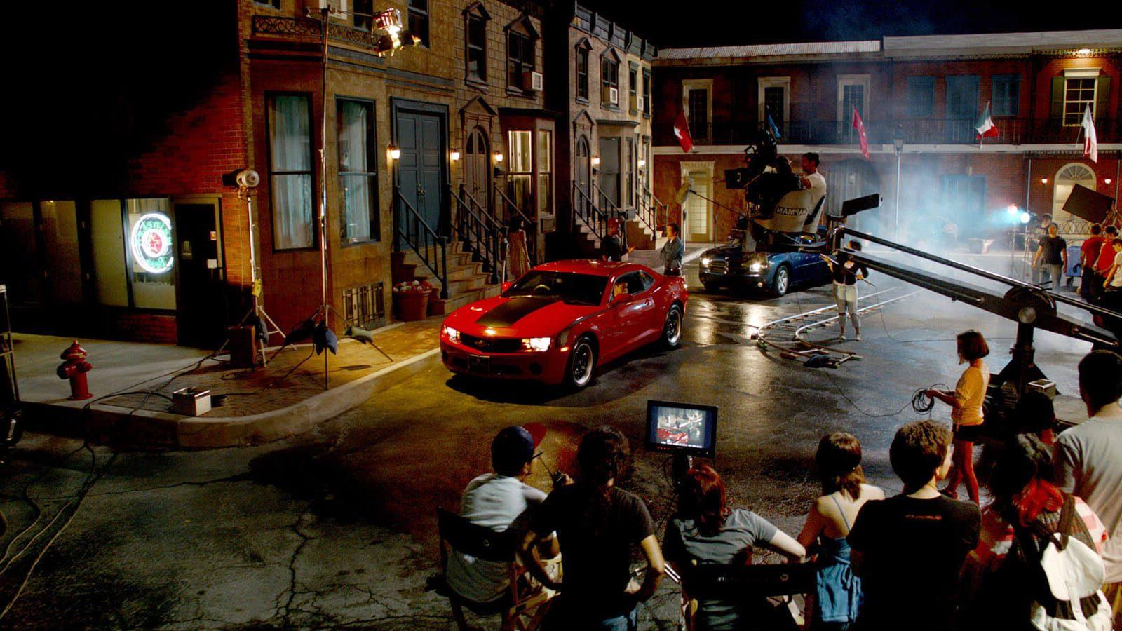 Full Sail University's Backlot in the evening filled with a full film production crew shooting a scene featuring two muscle cars, one red and one black.