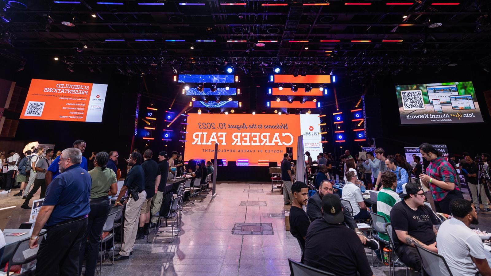 A bustling career fair with several booths set up. In the center, a giant LED screen welcomes guests to the Career Fair.