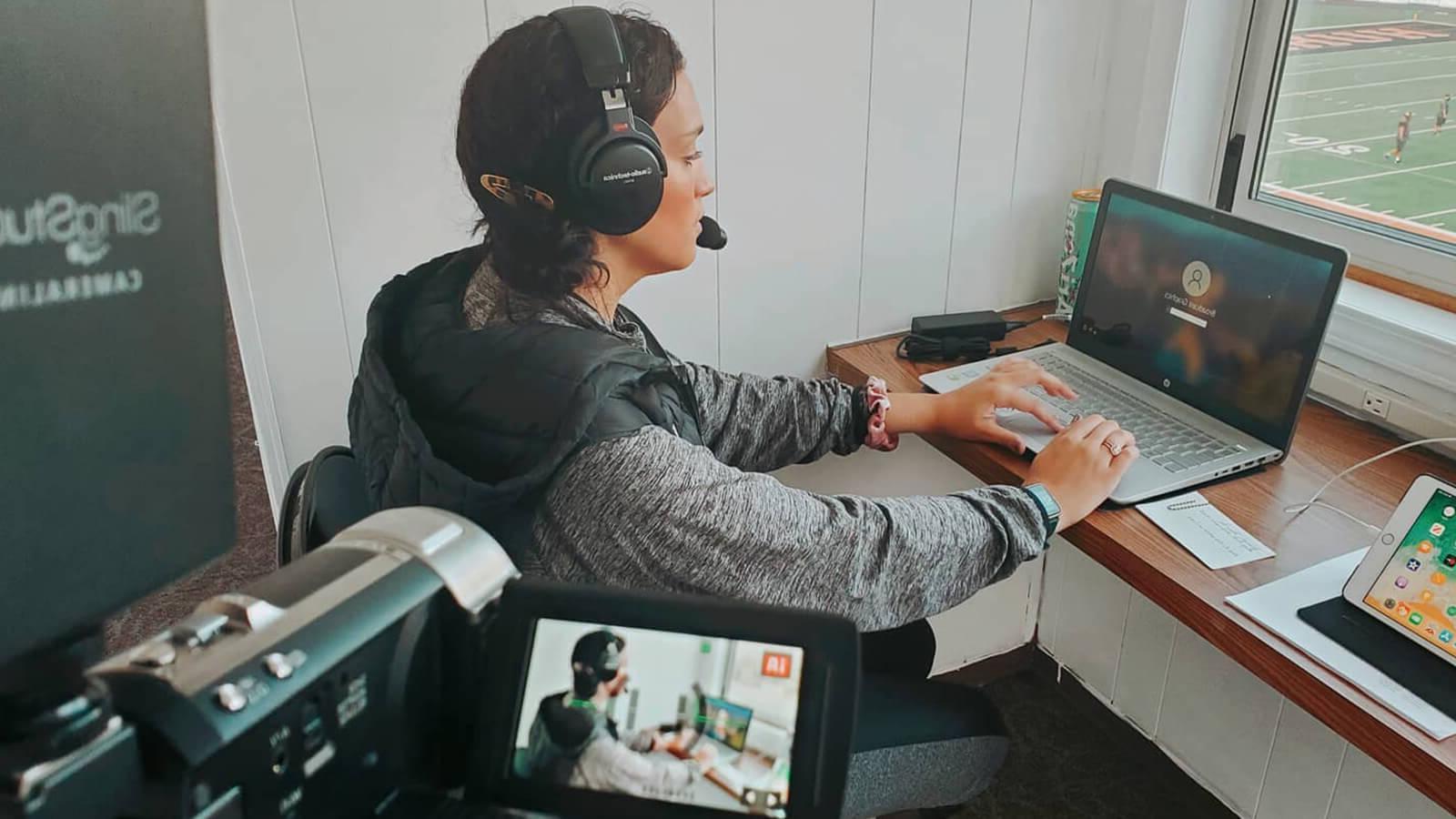 Grad Sydney Phillips, a woman with dark brown hair wearing a grey hooded sweatshirt and black over-ear headphones, is seated at a desk on her laptop in a press box overlooking athletes in uniform on a football field.