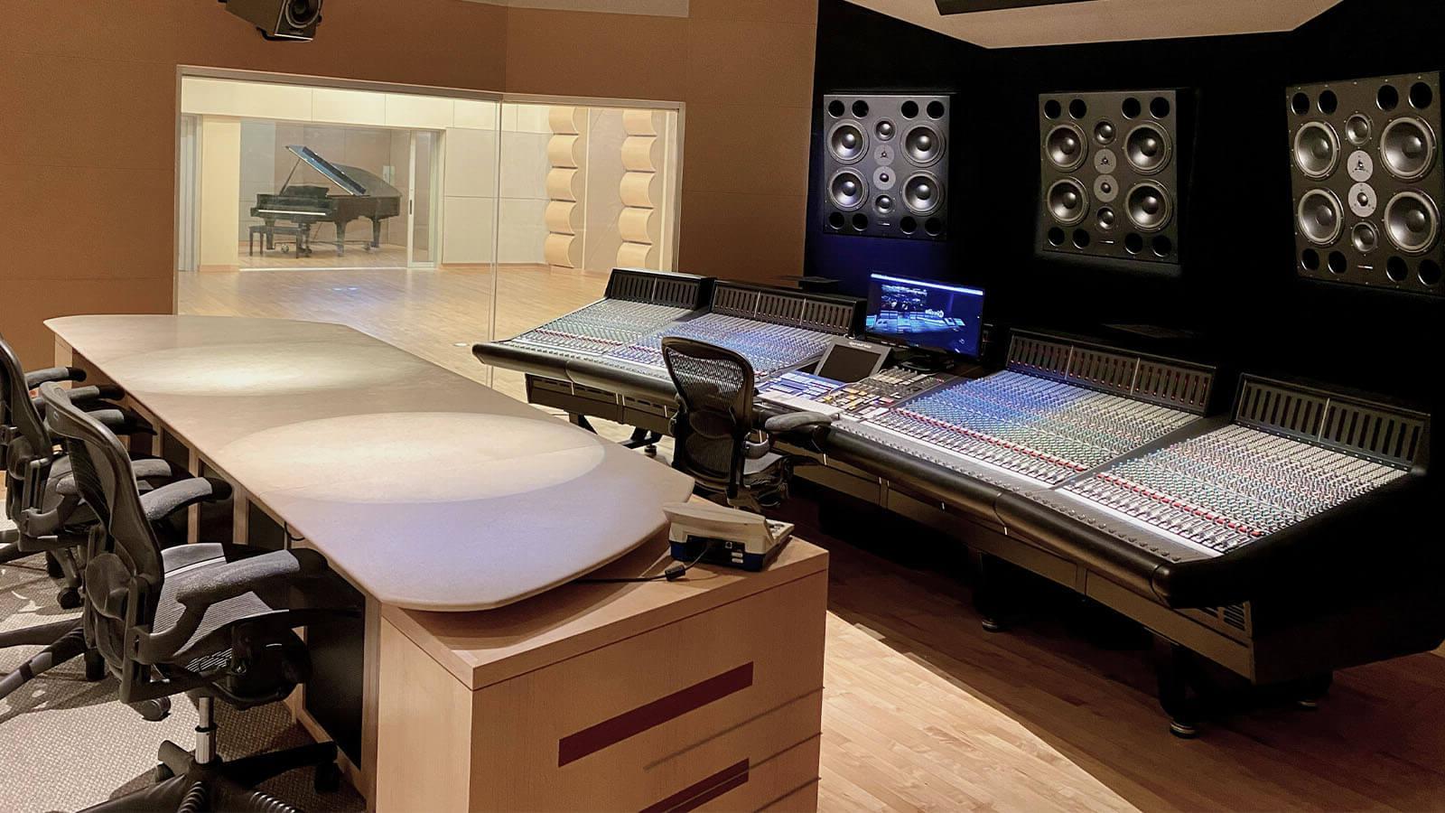 A conference table faces a wall with mixing boards and speakers; a recording area with a grand piano is in the background.