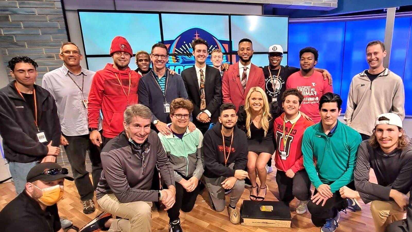 A group shot of several Full Sail students and instructors in front of a large screen on set at the Dan Patrick School of Sportscasting.