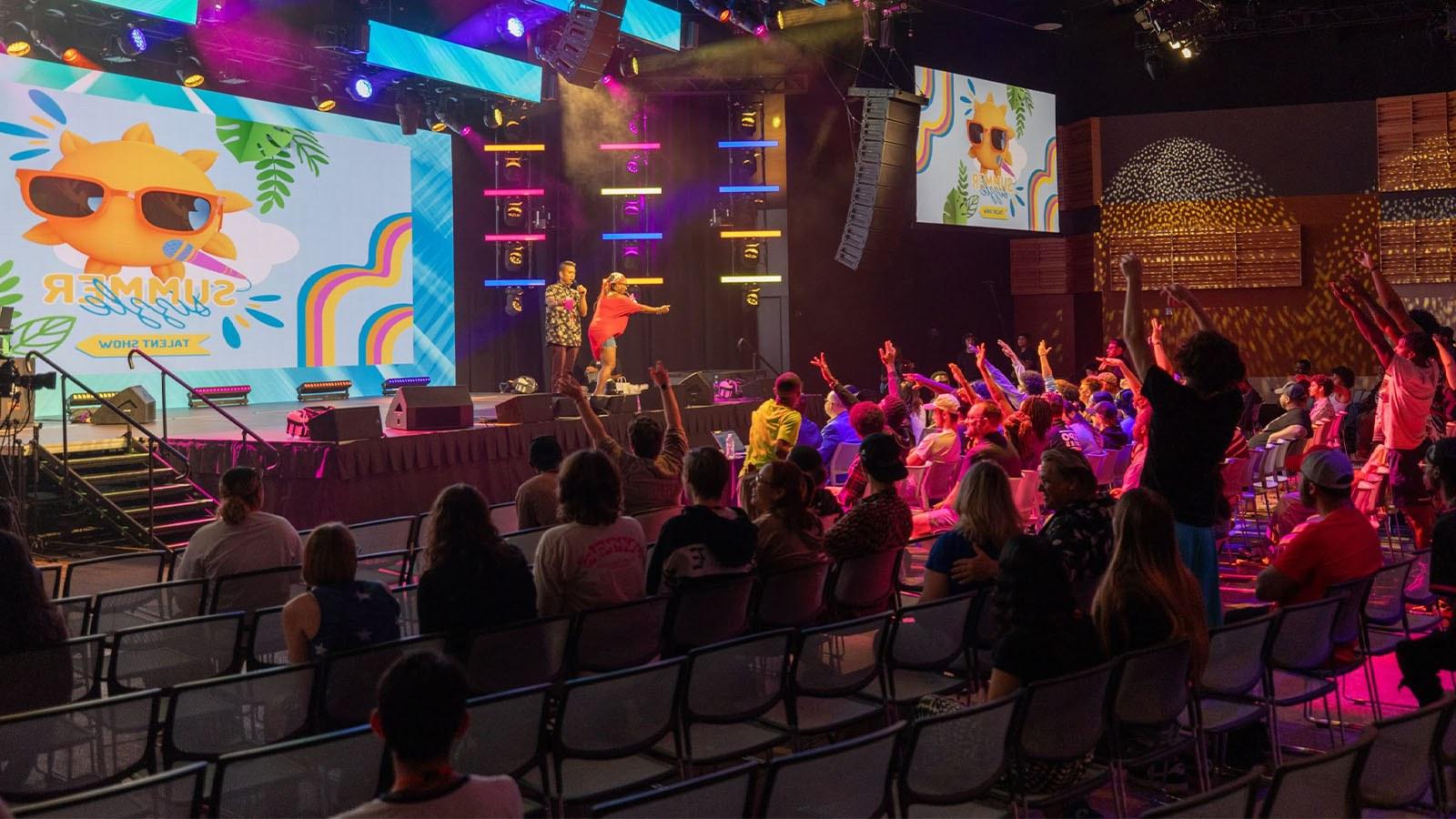 Several students raise their hands attentively at the Summer Sizzle as two staff members call on participants. The large room is colorfully lit up and looks very lively.