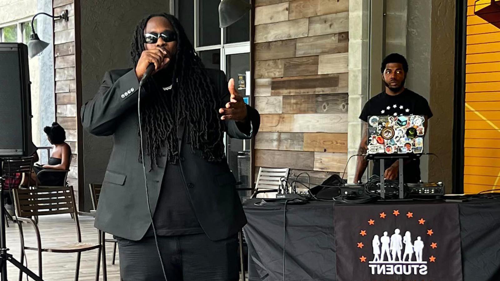 Guest speaker Souljah Bless, dressed in an all black outfit, speaks into a microphone on a patio stage. Behind him, a DJ stands at a booth that reads “Student Community Association.