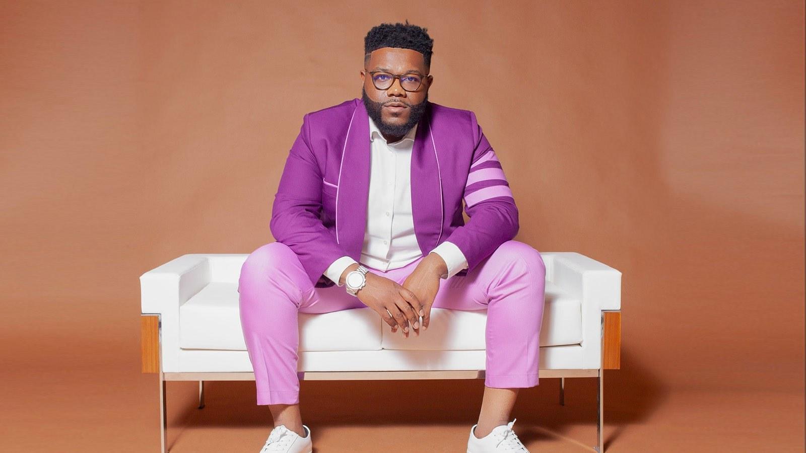 A man with black hair and a beard wearing glasses, a purple suit, and a white button down shirt while seated on a white couch against a brown background.