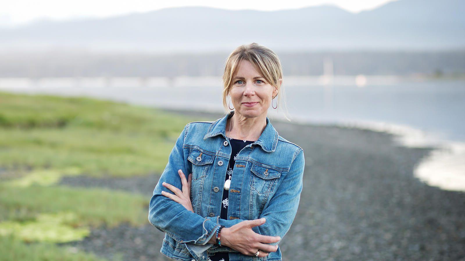 A blonde woman wearing a denim jacket over a navy top and black skirt stands with her arms crossed. She is outdoors in front of a river with mountains far in the background.