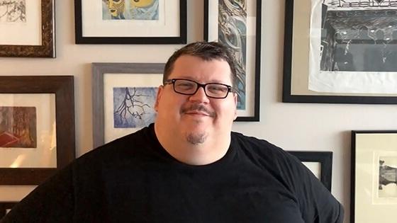 A smiling man wearing glasses and a black t-shirt sits in front of a wall covered with framed paintings.