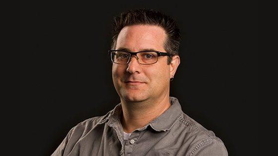 A man with short brown hair smiling at the camera while wearing black frame glasses and a gray button down against a black backdrop.