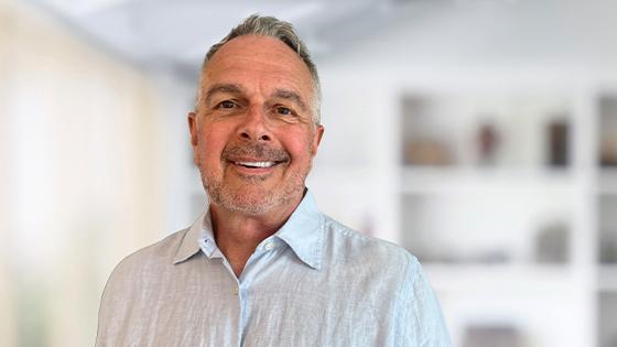 A person wearing a light blue button down while smiling against a white out-of-focus backdrop.