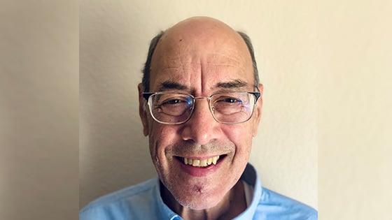 Dr. Carstens stands in front of a white wall wearing a blue shirt and glasses. He is smiling.