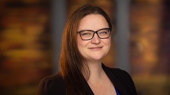 Rebecca Leis has brown hair and is wearing tortoiseshell glasses and a black blazer. She is smiling.