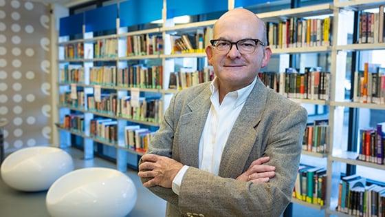 Rolando stands in a library with his arms crossed. He is smiling and wearing a suit.