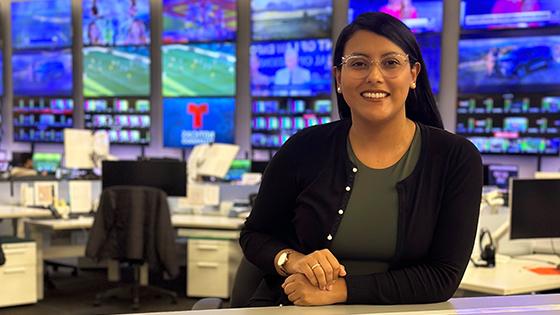 Priscilla sits at a desk in a live news television studio. 她戴着眼镜，面带微笑.