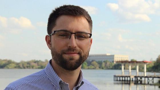 A man wearing glasses and a button-up shirt stands in front of a pier.