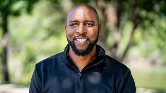 Full Sail grad Sidney Walker in the foreground smiling while wearing a black windbreaker with lush greenery behind him.