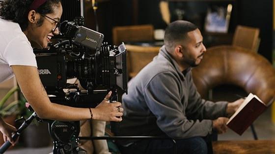 Behind the scenes on a student film set, one student sits in a chair with a notebook while another student works behind a film camera