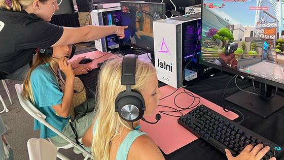 Two young girls are seated at desktop computers playing 'Minecraft' while a woman wearing a Super Girl Gamer Pro shirt assists them.