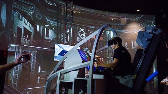 A student wearing a VR headset sits in a space shuttle landing simulator. A large screen displaying a warehouse is in front of them.