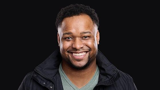 James Lewis smiles at the camera. He is wearing a black jacket and green v-neck t-shirt.