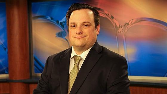 Dan Hanson sits at a news desk in a television studio. He is smiling and wearing a black suit with a yellow tie.