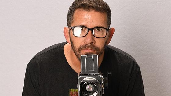 Sebastian Krys crouches behind a video camera. He is wearing black-rimmed eyeglasses and a black t-shirt with a logo on it.