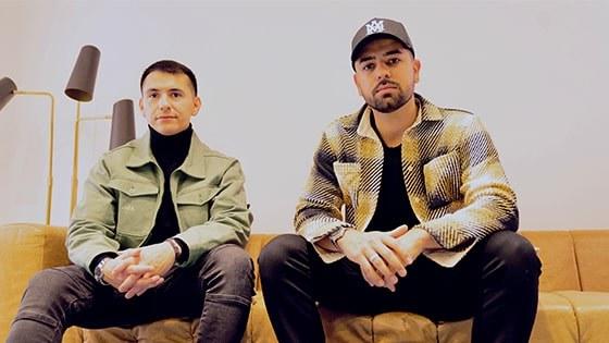 Two men seated side by side on a light brown leather couch against a white backdrop.