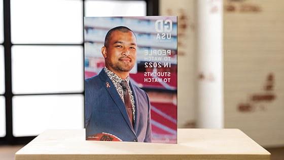 A Graphic Design USA magazine stands upright on a desk in a backlit room. On the cover, a man wearing a blue suit with a pink ribbon pin and holding an NFL football smiles for the camera. The cover reads, People to Watch in 2022, Students to Watch.