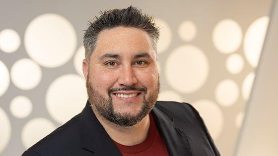 Michael La Plante smiles at the camera. He is wearing a red t-shirt with a black blazer and standing in front of a white backdrop.